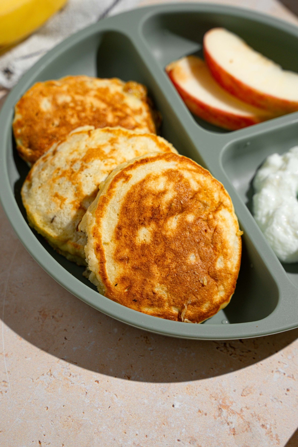 A kids plate with three 3 ingredient banana pancakes with a side of sliced apples and cottage cheese. The plate is on a tan counter with a white striped napkin and bananas.
