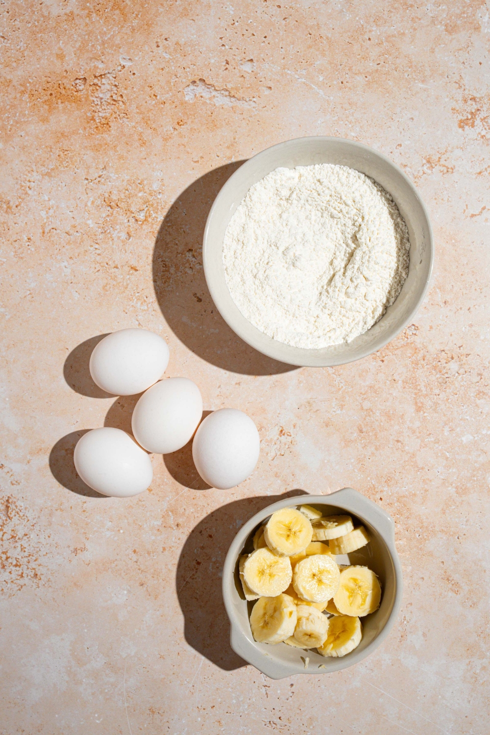 An overhead shot of three ingredients to make banana pancakes including flour, eggs, and sliced bananas.
