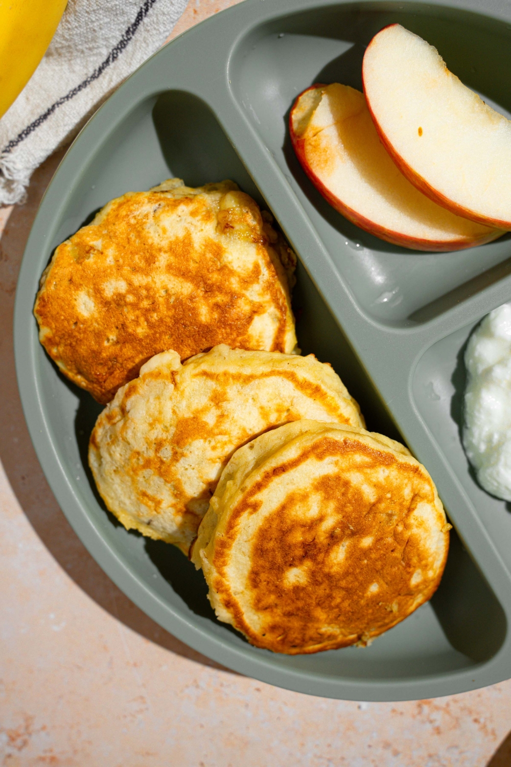 A kids plate with three 3 ingredient banana pancakes with a side of sliced apples and cottage cheese. The plate is on a tan counter.