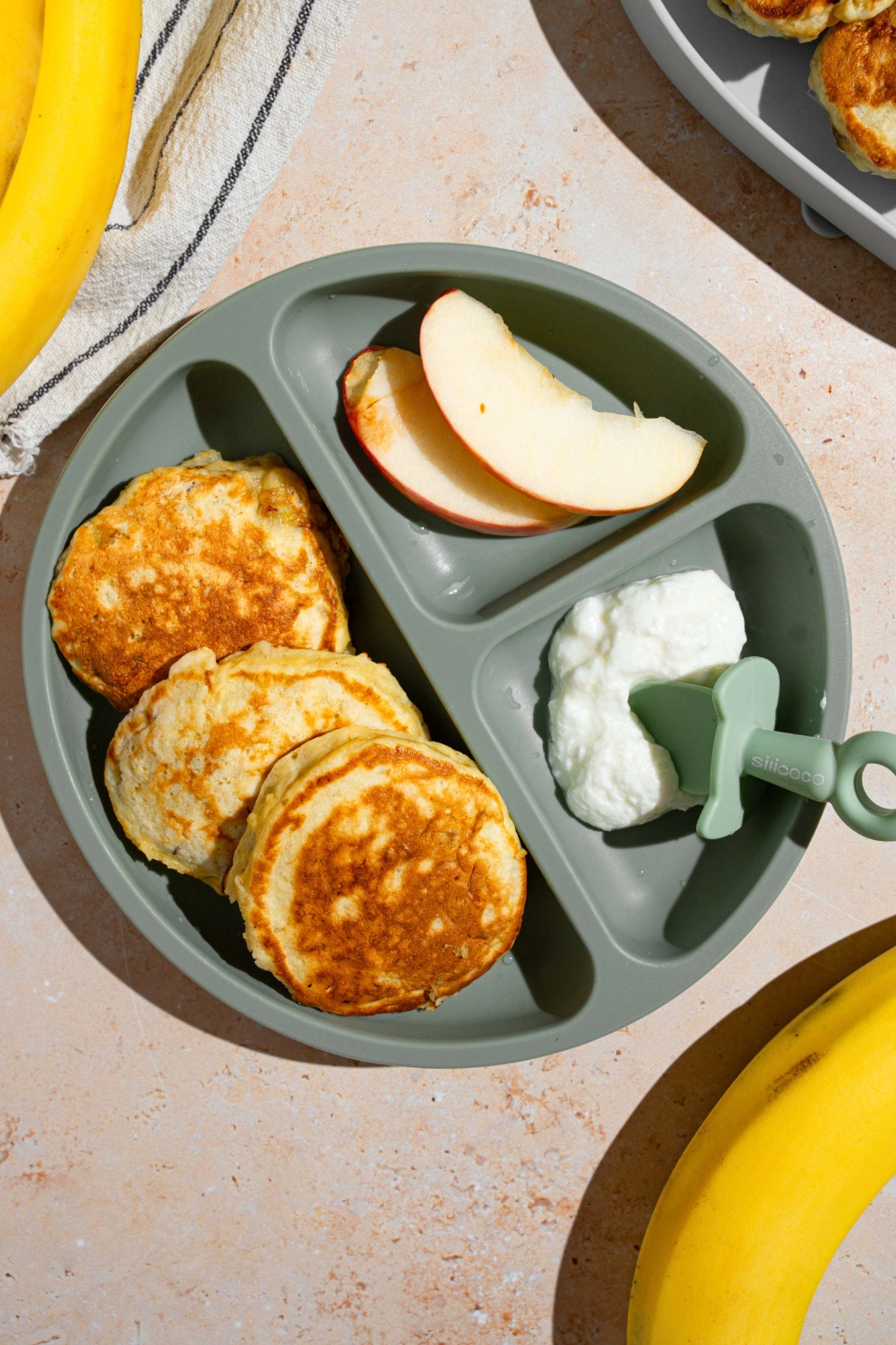A kids plate with three 3 ingredient banana pancakes with a side of sliced apples and cottage cheese. The plate is on a tan counter with a white striped napkin and bananas.