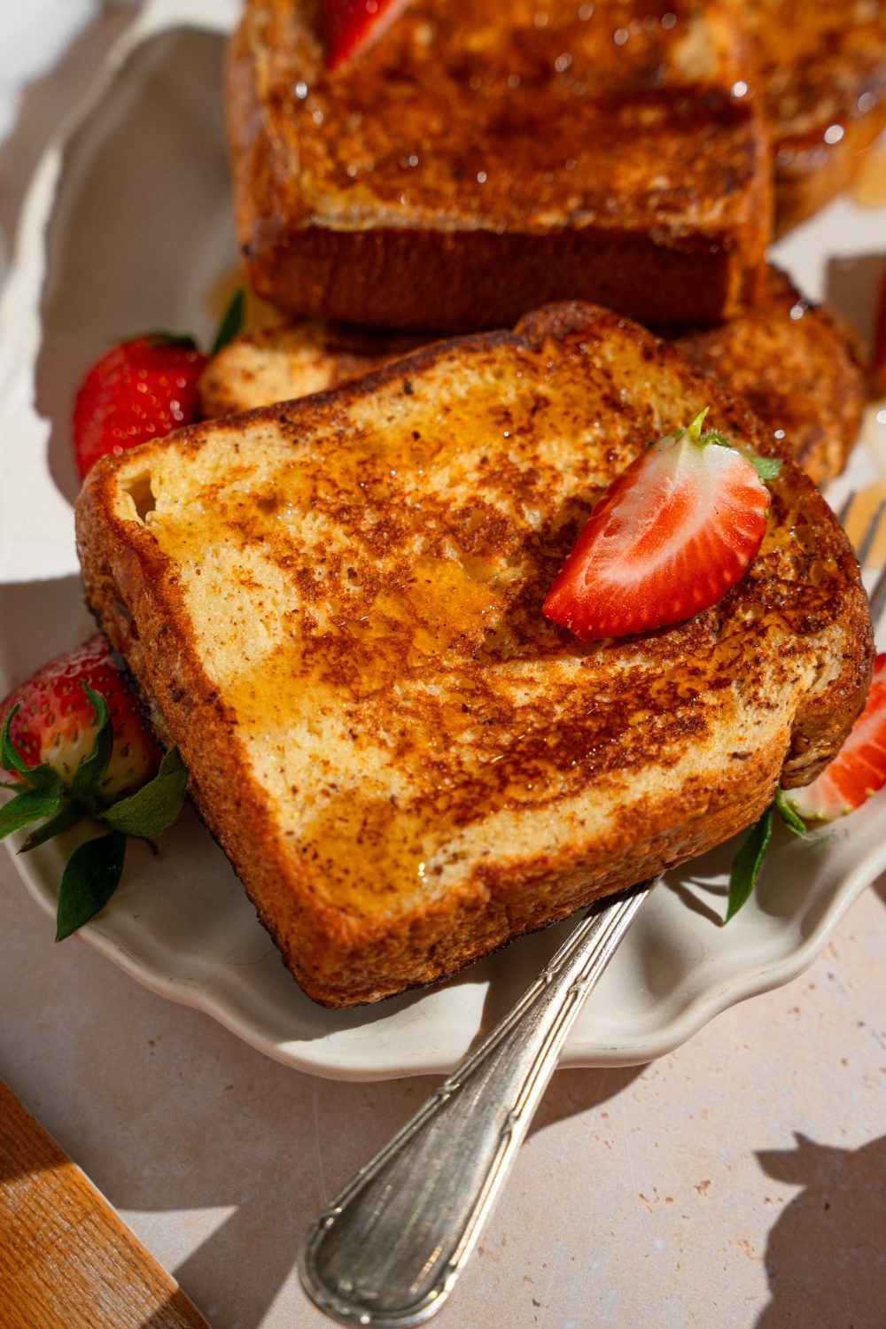 A white platter with slices of French toast drizzled with maple syrup and garnished with sliced strawberries. There is a fork on the plate.