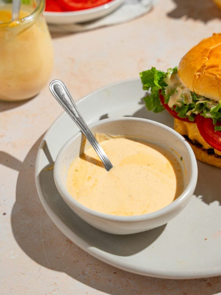 A bowl of homemade Shake Shack sauce with a spoon in the bowl. The bowl is on a white plate with a Shake Shack burger. The plate is on a tan counter with an additional jar of sauce.