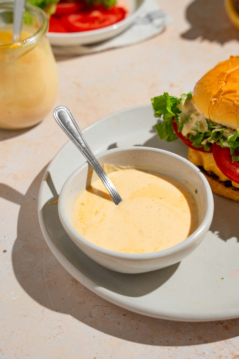 A bowl of homemade Shake Shack sauce with a spoon in the bowl. The bowl is on a white plate with a Shake Shack burger. The plate is on a tan counter with an additional jar of sauce.