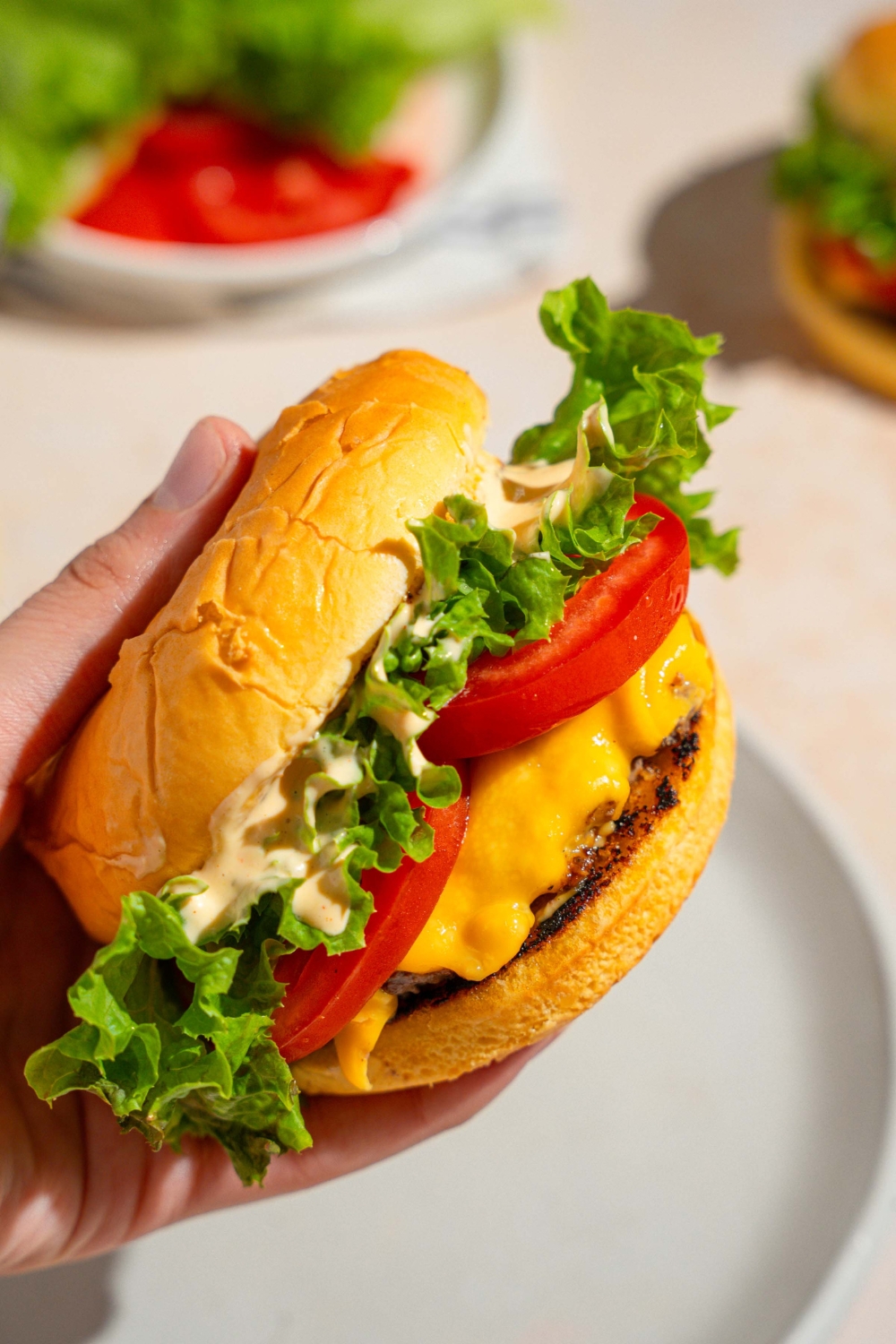 A close up of a hand holding a homemade Shake Shack burger with Shake Shack sauce. There is a white plate on a tan counter blurred in the background.