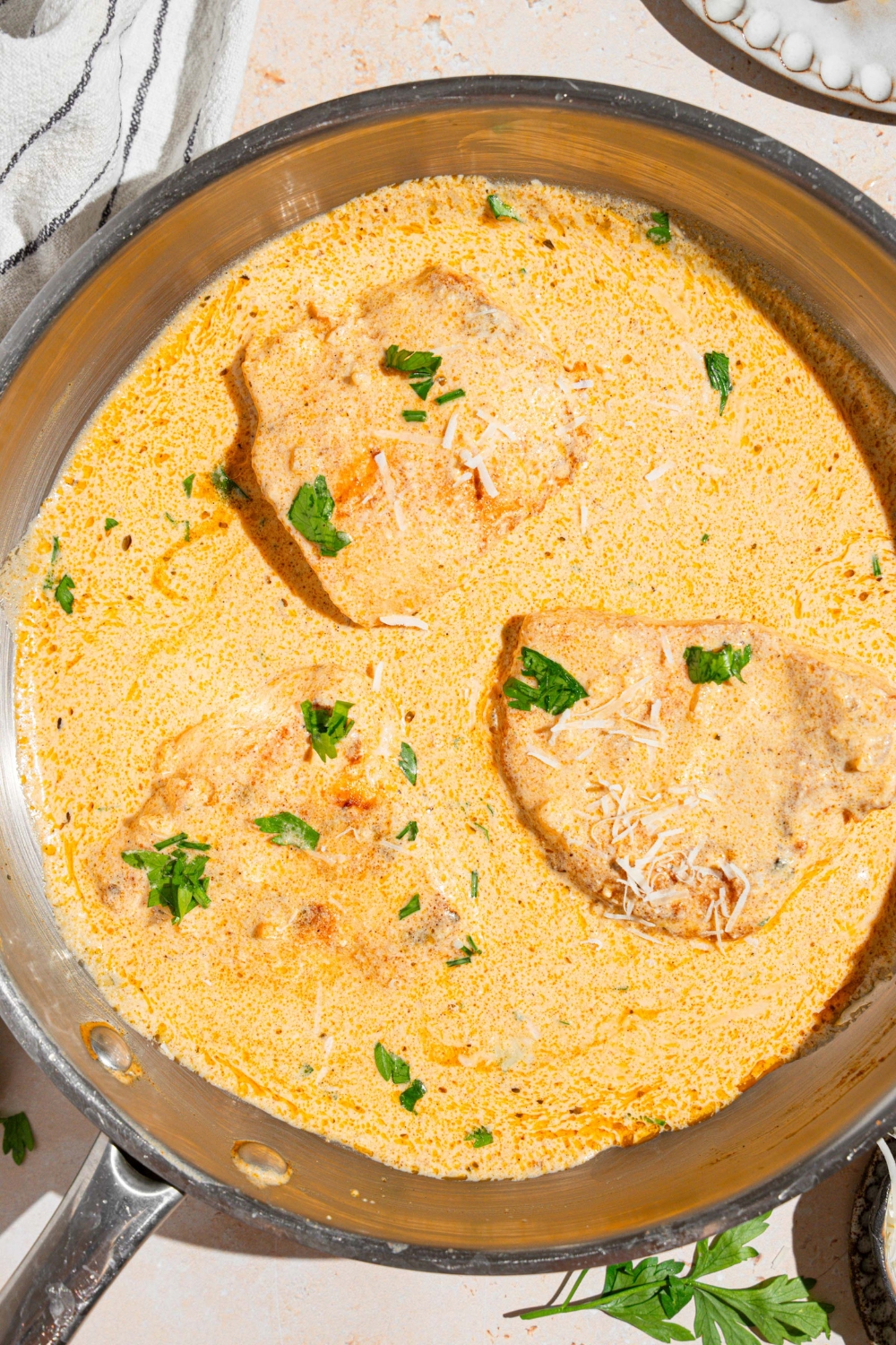 A skillet with chicken breasts cooked in cajun alfredo sauce garnished with fresh parlsey and grated cheese. The skillet is on a tan counter with a white striped napkin.