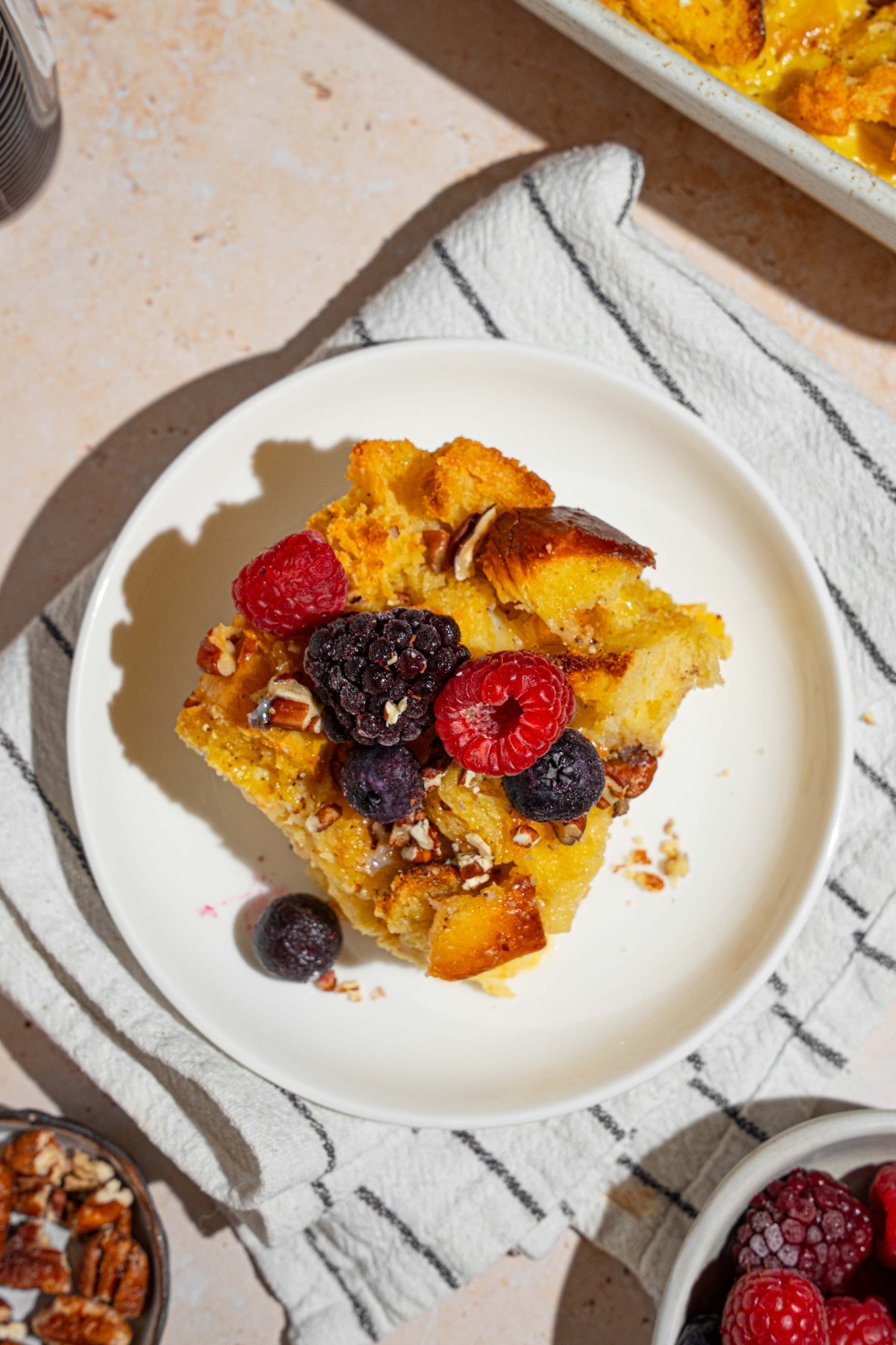 A white plate with a slice of challah french toast topped with mixed berries. The plate is on a tan counter with a white striped napkin with a bowl of berries and plate of pecans.