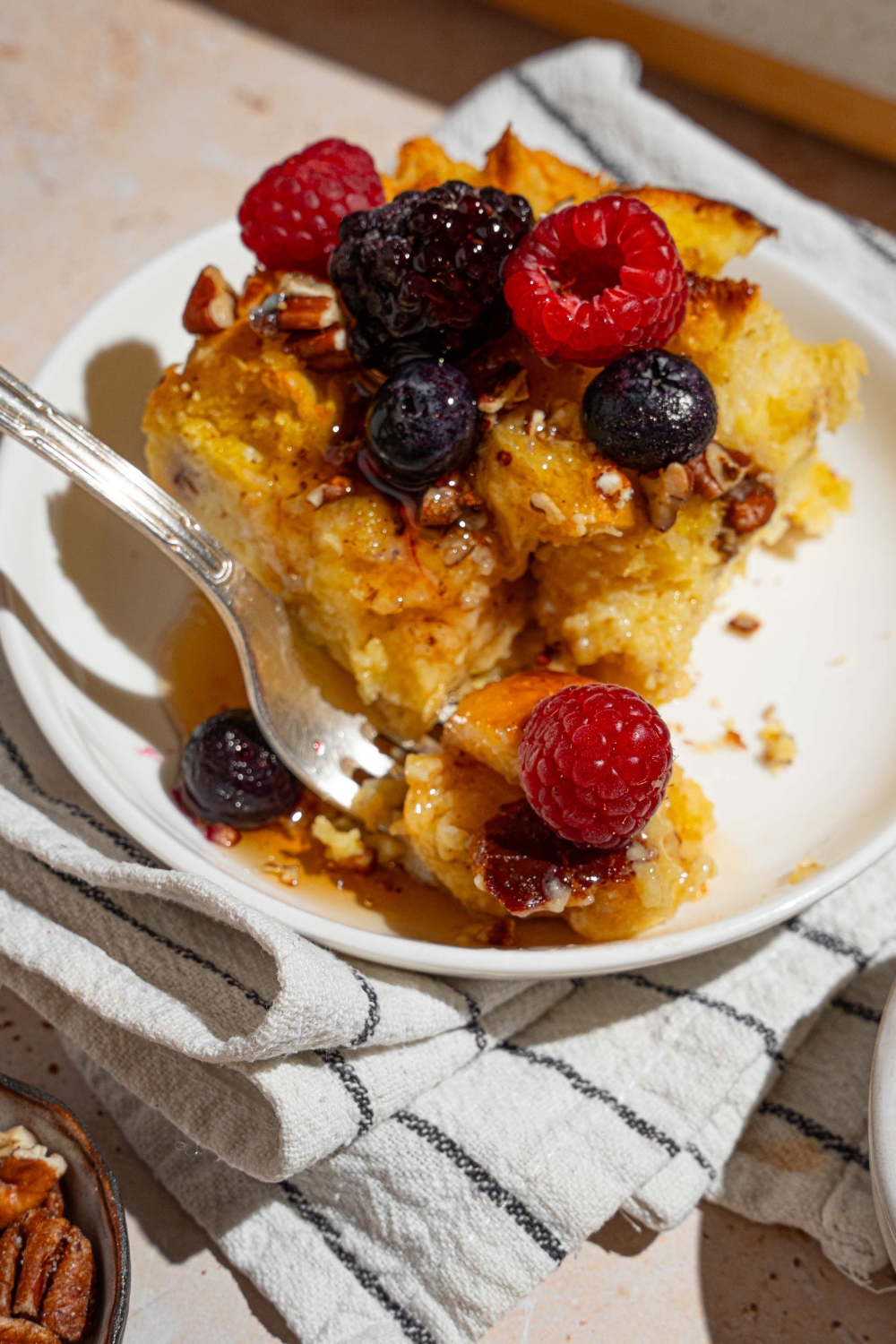 A white plate with a slice of challah french toast topped with mixed berries drizzled with syrup. A fork is on the plate with a bite of french toast. The plate is on a tan counter with a white striped napkin.