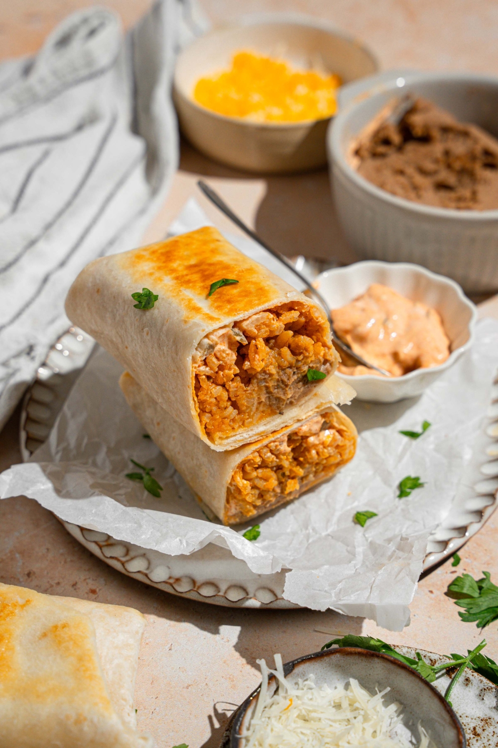 A cheesy bean and rice burrito sliced in half with each half stacked on the other. The burrito is on a plate lined with parchment paper with a small side of creamy jalapeno sauce with a spoon. The plate is garnished with fresh parsley. The plate is on a tan counter with small bowls of refried beans and shredded cheese.