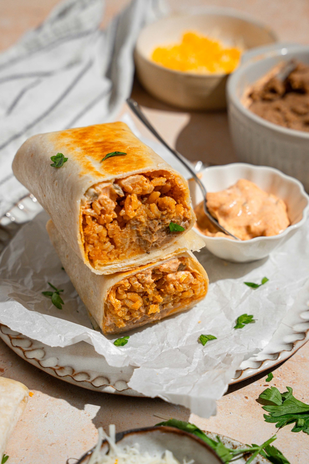 A cheesy bean and rice burrito sliced in half with each half stacked on the other. The burrito is on a plate lined with parchment paper with a small side of creamy jalapeno sauce with a spoon. The plate is garnished with fresh parsley. The plate is on a tan counter with small bowls of refried beans and shredded cheese.