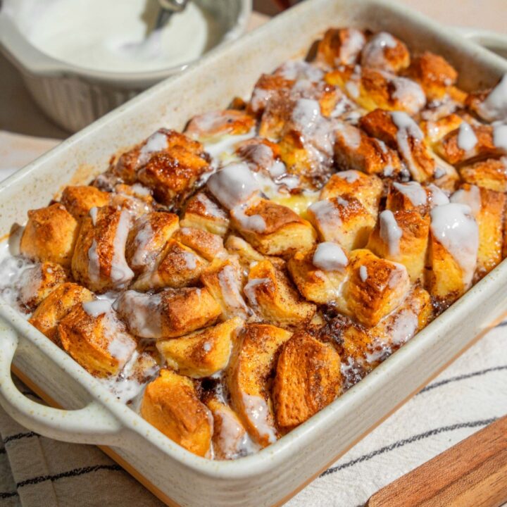 A baking dish with cinnamon roll french toast drizzled with icing. The dish is on a tan counter on a white striped napkin. The dish is on a tan counter with a small bowl of icing.