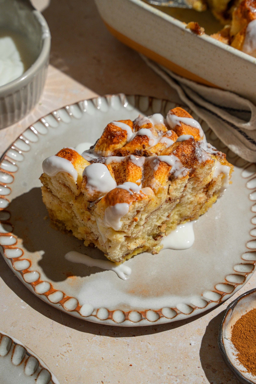 A ceramic plate with a slice of cinnamon roll french toast drizzled with icing. The plate is on a tan counter with a baking dish of french toast.