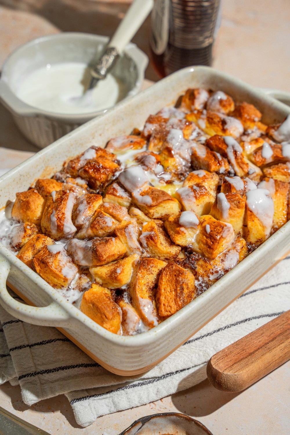 A baking dish with cinnamon roll french toast drizzled with icing. The dish is on a tan counter on a white striped napkin. The dish is on a tan counter with a small bowl of icing.