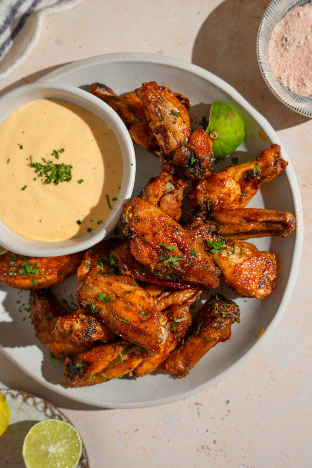 A white plate with copycat Wingstop Louisiana rub wings garnished with fresh parsley, a lime wedge, and small bowl of wing sauce. The plate is on a tan counter with a small plate of rub.