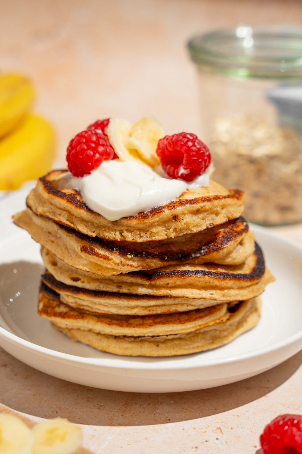 A white plate with a stack of cottage cheese banana pancakes topped with whipped cream, sliced banana, and raspberries. The plate is on a tan counter with bananas and a jar of oats.