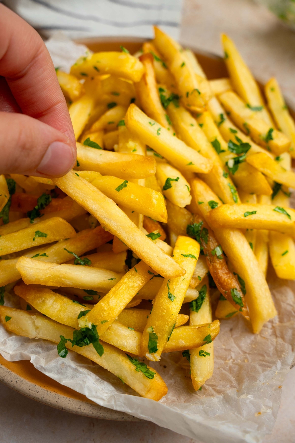 Garlic fries garnished with fresh parsley served on a plate lined with parchment paper. A hand is taking fries from the plate.