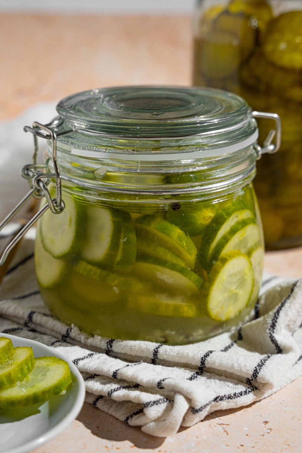 A jar with quick pickles in juices. The jar is on a white striped napkin on a tan counter with a small plate of sliced pickles.