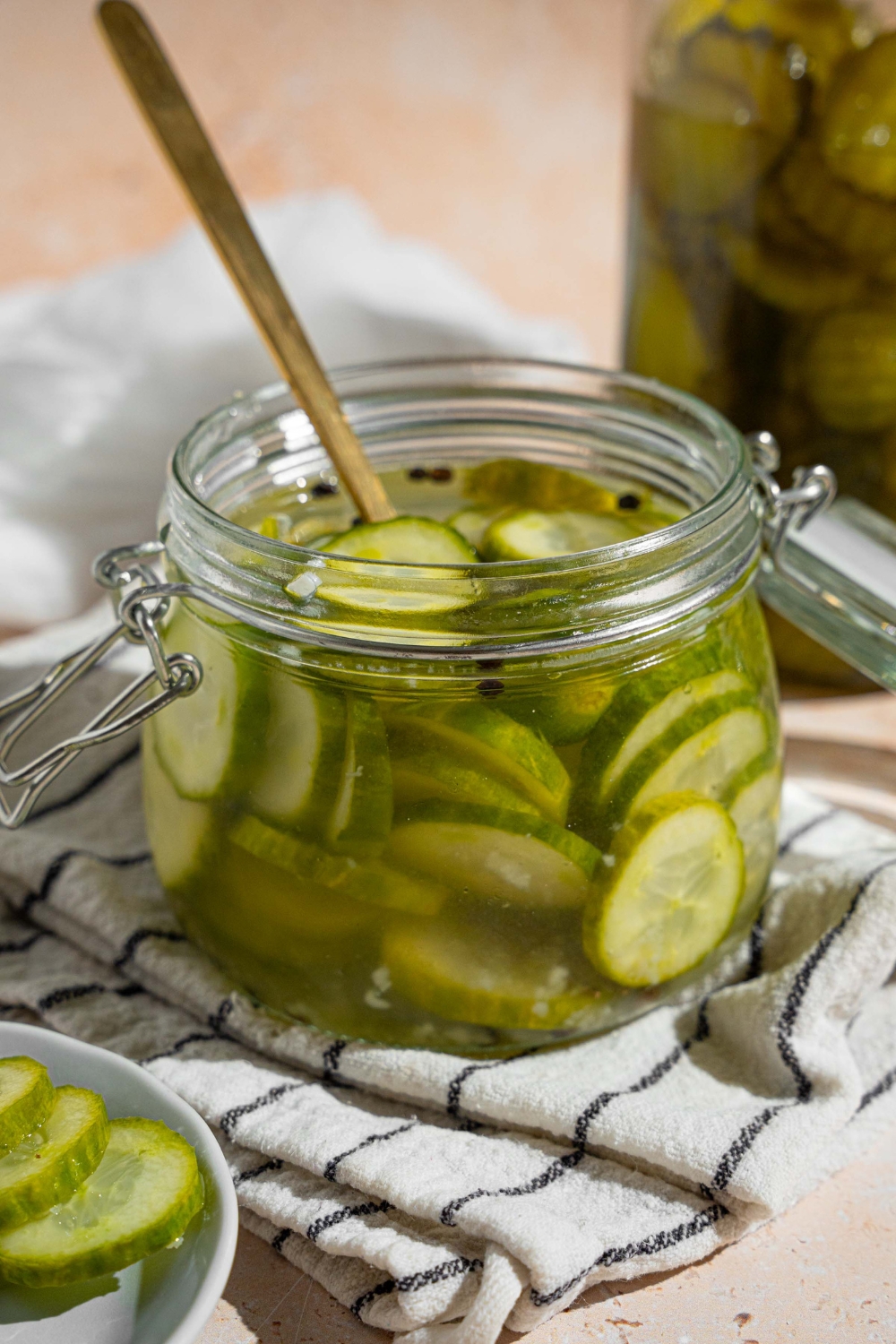 A jar with quick pickles in juices. There is a fork in the jar. The jar is on a white striped napkin on a tan counter with a small plate of sliced pickles.