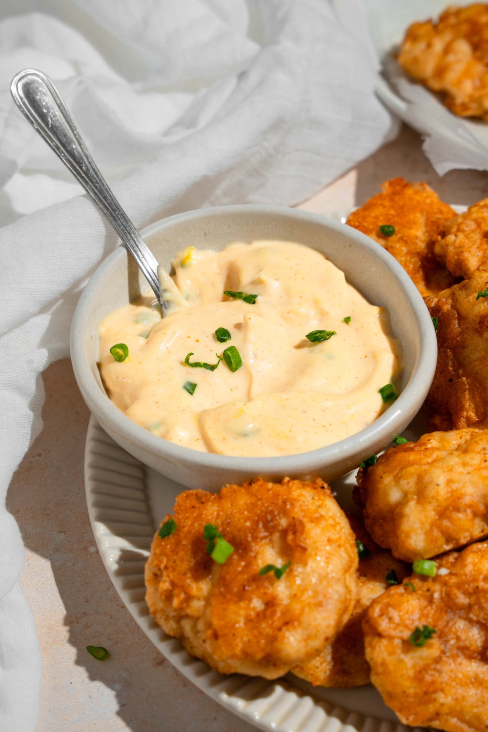 A ramekin of remoulade sauce garnished with chopped chives and served with a spoon. The bowl is on a plate of chicken nuggets on a tan counter with a white cloth napkin.