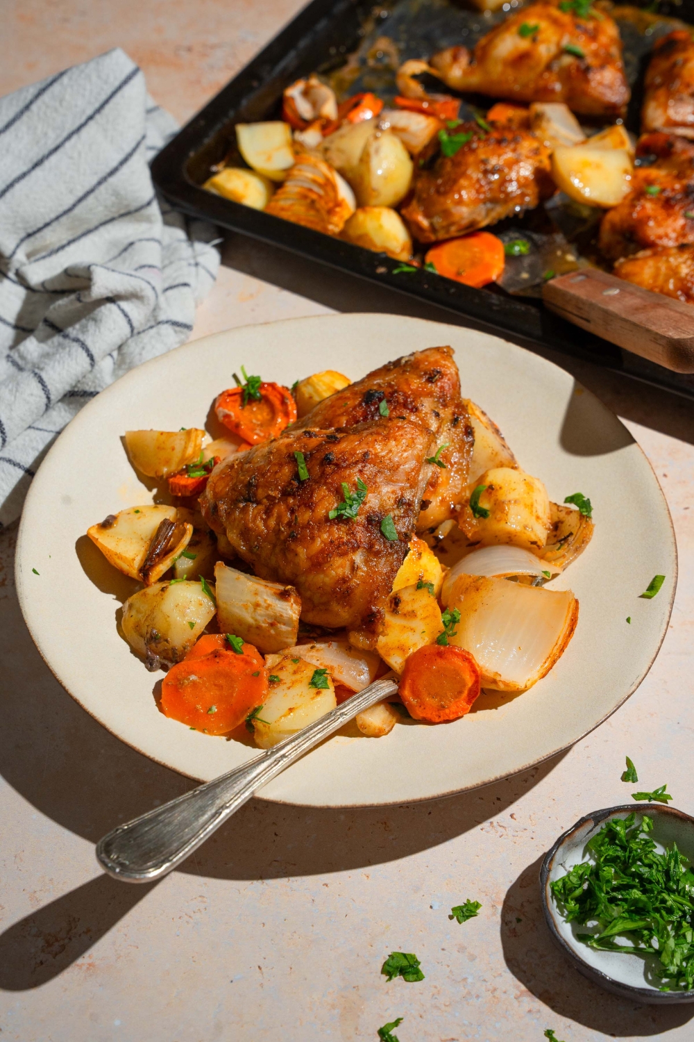A white plate with a roasted chicken thigh served over roasted carrots, potatoes, and onions garnished with fresh parsley. There is a fork in the plate. The plate is on a tan counter with a baking sheet of chicken and vegetables and a white striped napkin.