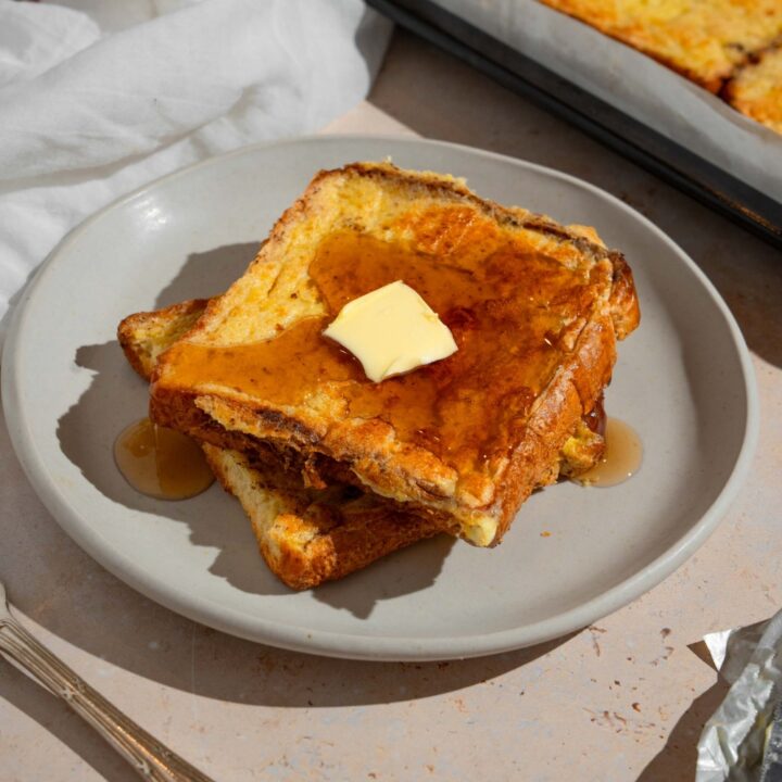 A plate with two slices of sheet pan french toast topped with butter and drizzled with syrup. The plate is on a tan counter with a fork and sheet pan of french toast.