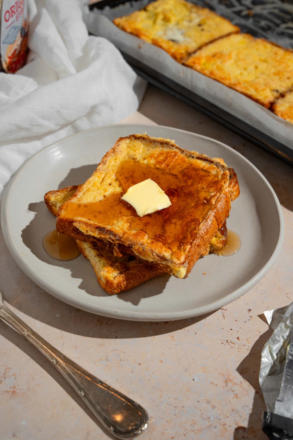 A plate with two slices of sheet pan french toast topped with butter and drizzled with syrup. The plate is on a tan counter with a fork and sheet pan of french toast.
