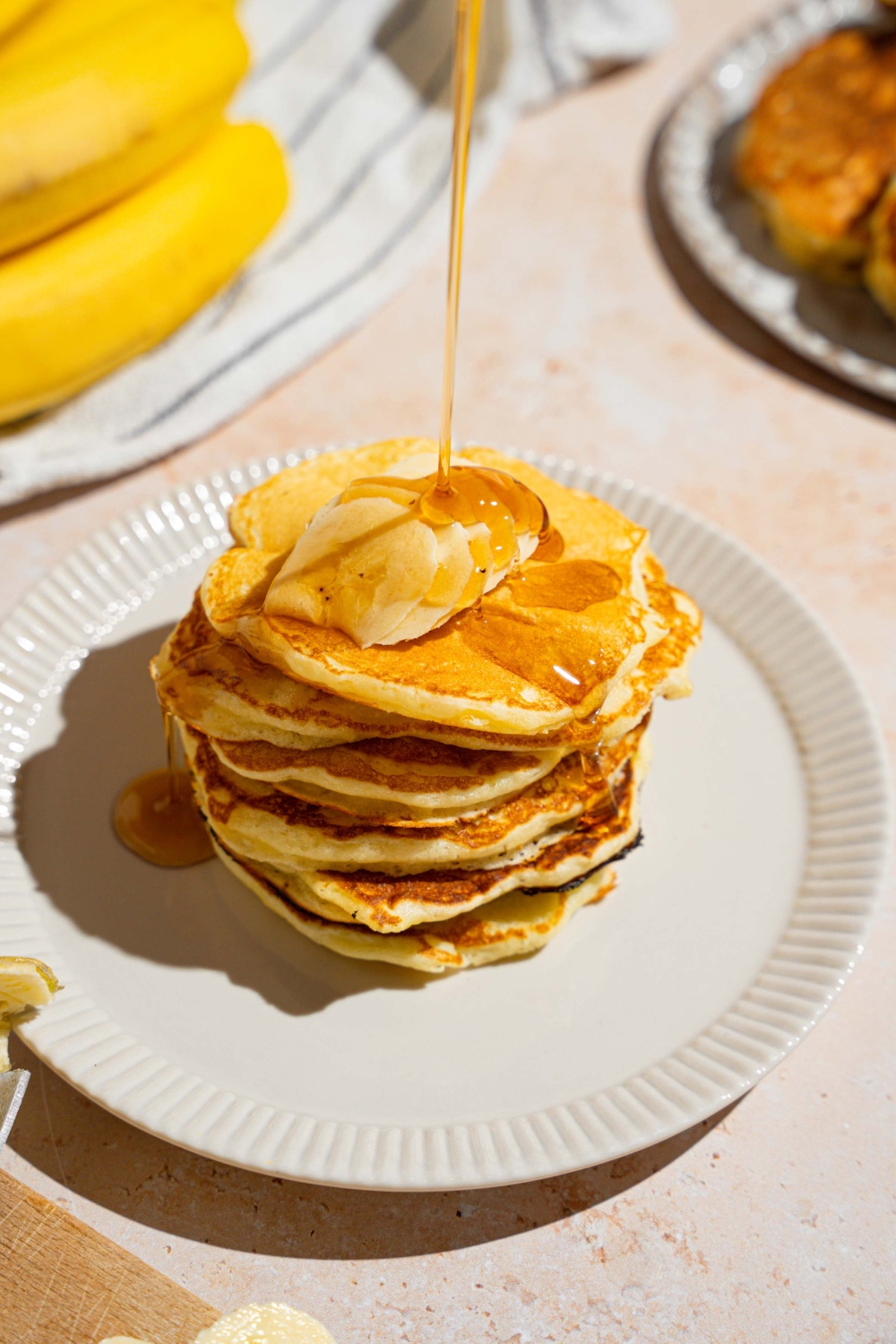 A stack of pancakes topped with butter. The pancakes are on a white plate with syrup being poured on the stack. The plate is on a tan counter with a white striped napkin and bananas.