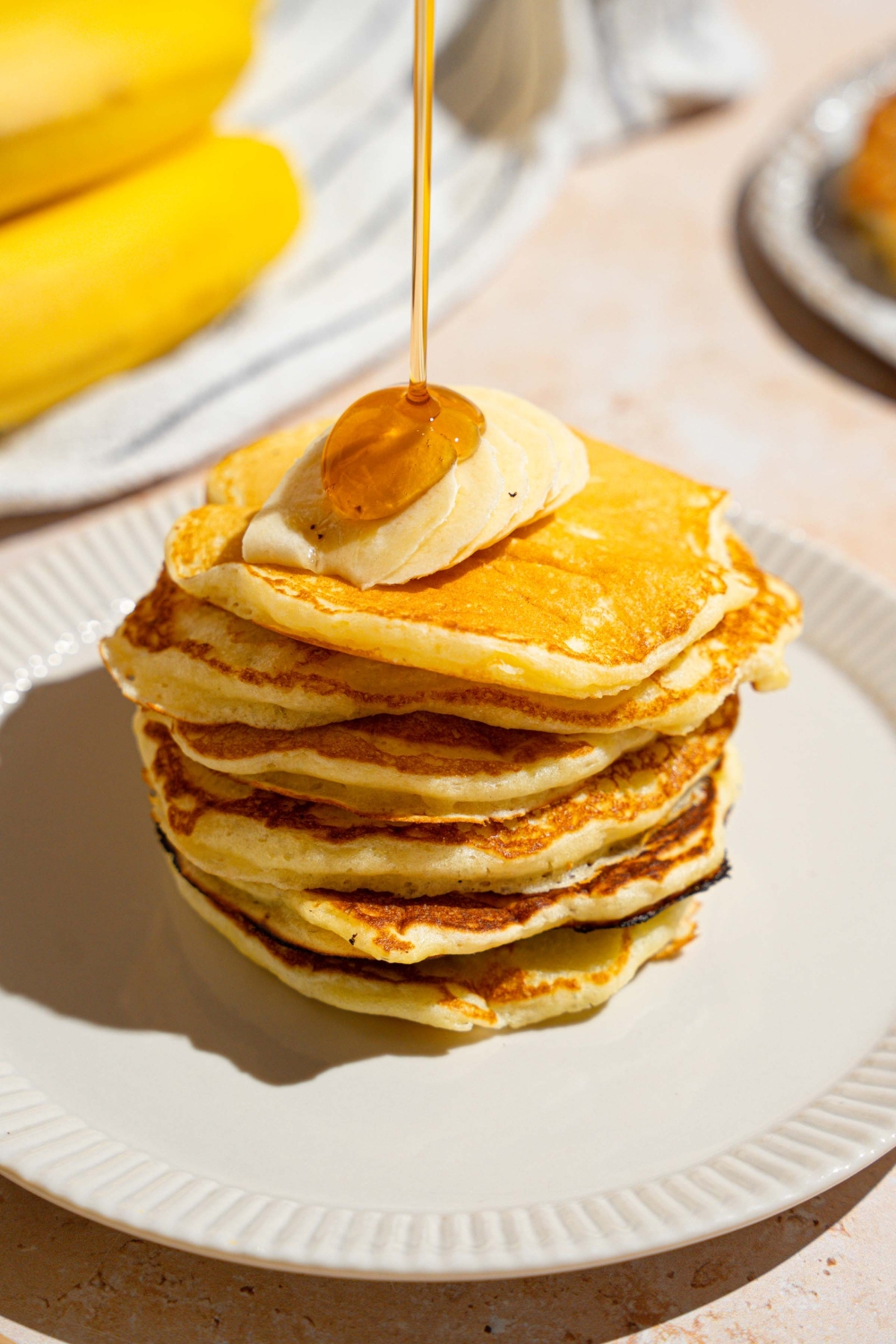 A stack of pancakes topped with sliced banana. The pancakes are on a white plate with syrup being poured on the stack. The plate is on a tan counter with a white striped napkin and bananas.