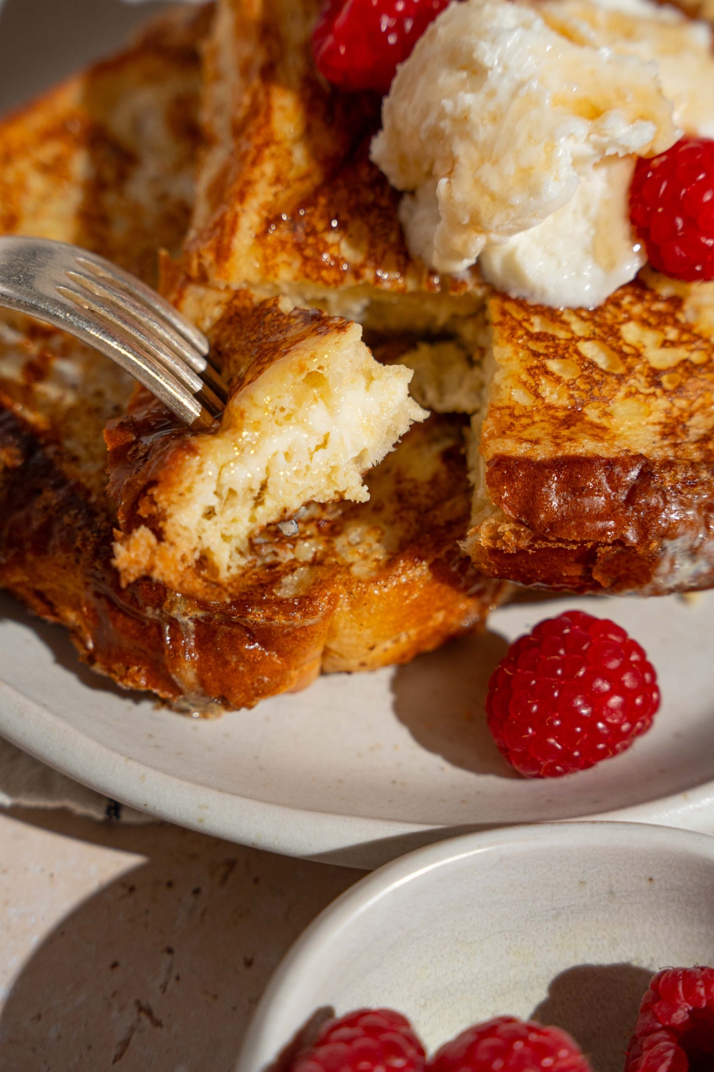 A white plate with stuffed french toast topped with whipped cream cheese and raspberries. There is a fork taking a bite of french toast.