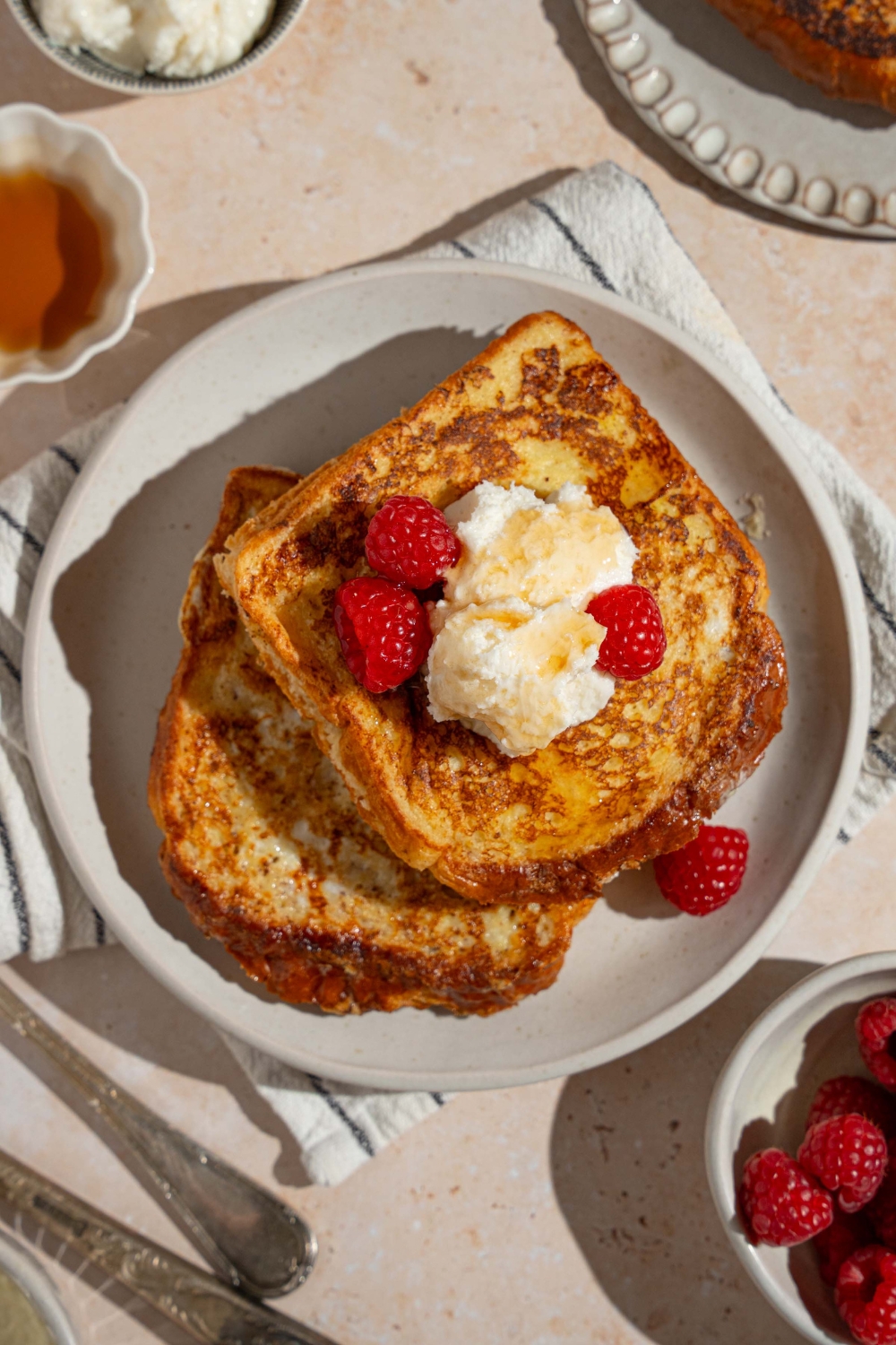 A white plate with stuffed french toast topped with whipped cream cheese and raspberries drizzled with syrup. The plate is on a tan counter with a white striped napkin, bowl of syrup, and bowl of raspberries.