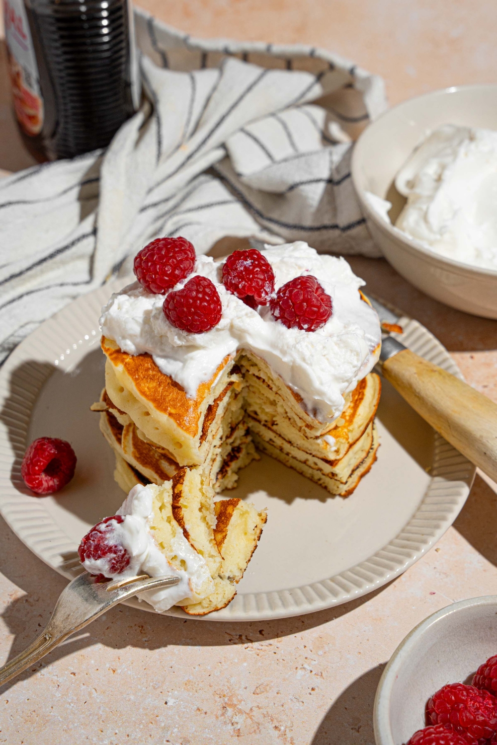 A white plate with a stack of sweet cream fluffy pancakes topped with whipped cream and raspberries. There is a fork taking a bite from the stack. The plate is on a tan counter with a white striped napkin and bowl of whipped cream.
