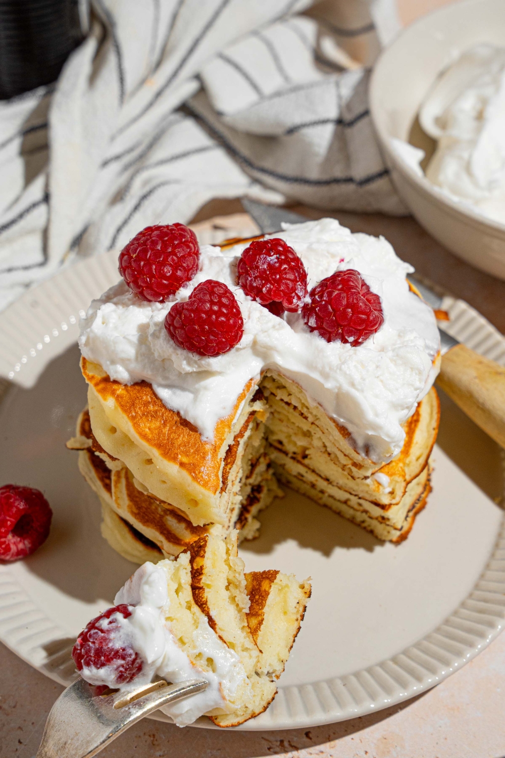 A white plate with a stack of sweet cream fluffy pancakes topped with whipped cream and raspberries. There is a fork taking a bite from the stack. The plate is on a tan counter with a white striped napkin and bowl of whipped cream.