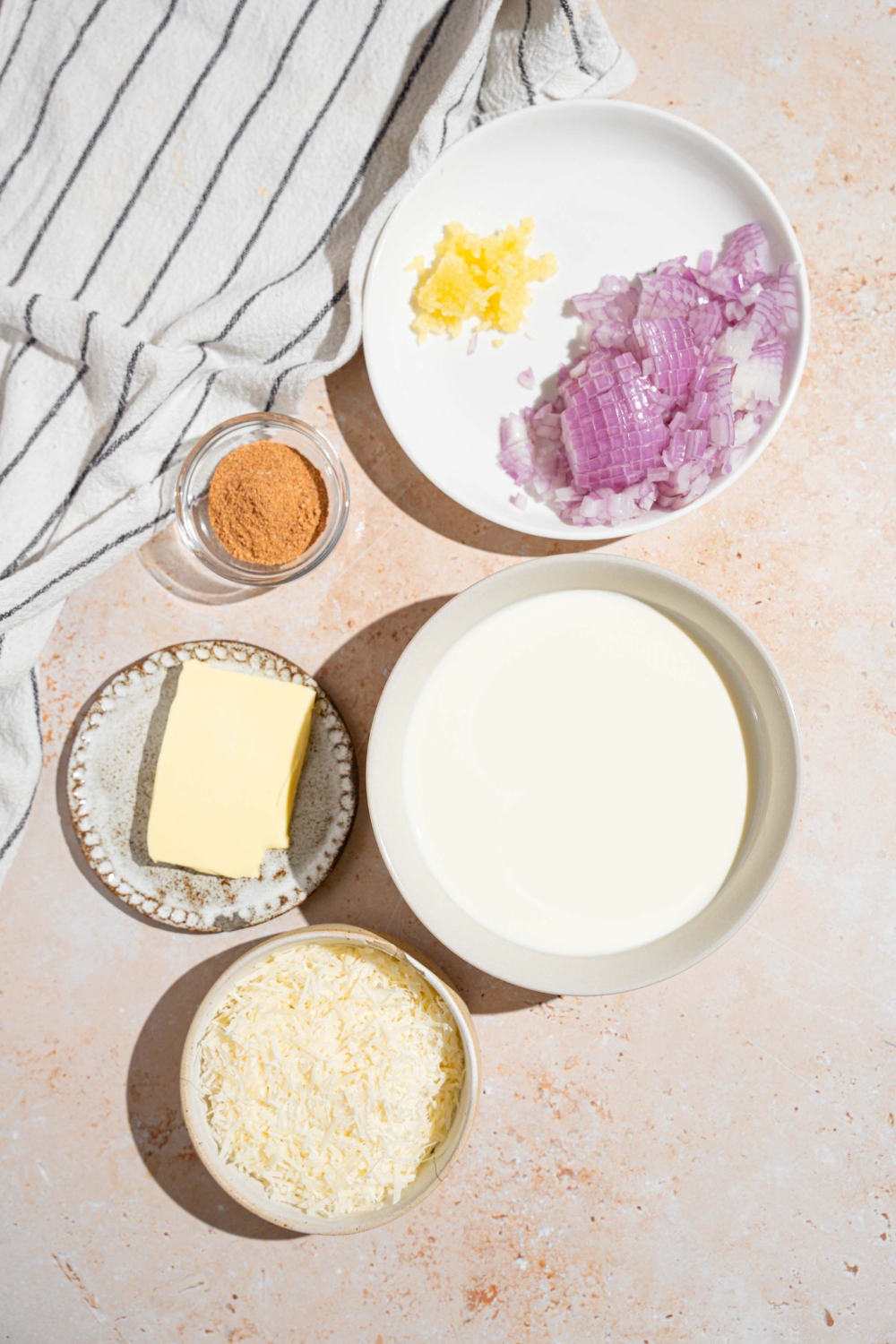 An overhead shot of several bowls in various sizes containing ingredients to make cajun alfredo sauce including butter, shallots, garlic, heavy cream, grated cheese, cajun seasoning, and more.