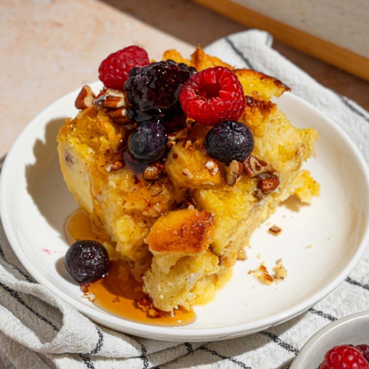 A white plate with a slice of challah french toast topped with mixed berries drizzled with syrup. The plate is on a tan counter with a white striped napkin with a bowl of berries and baking dish of french toast.