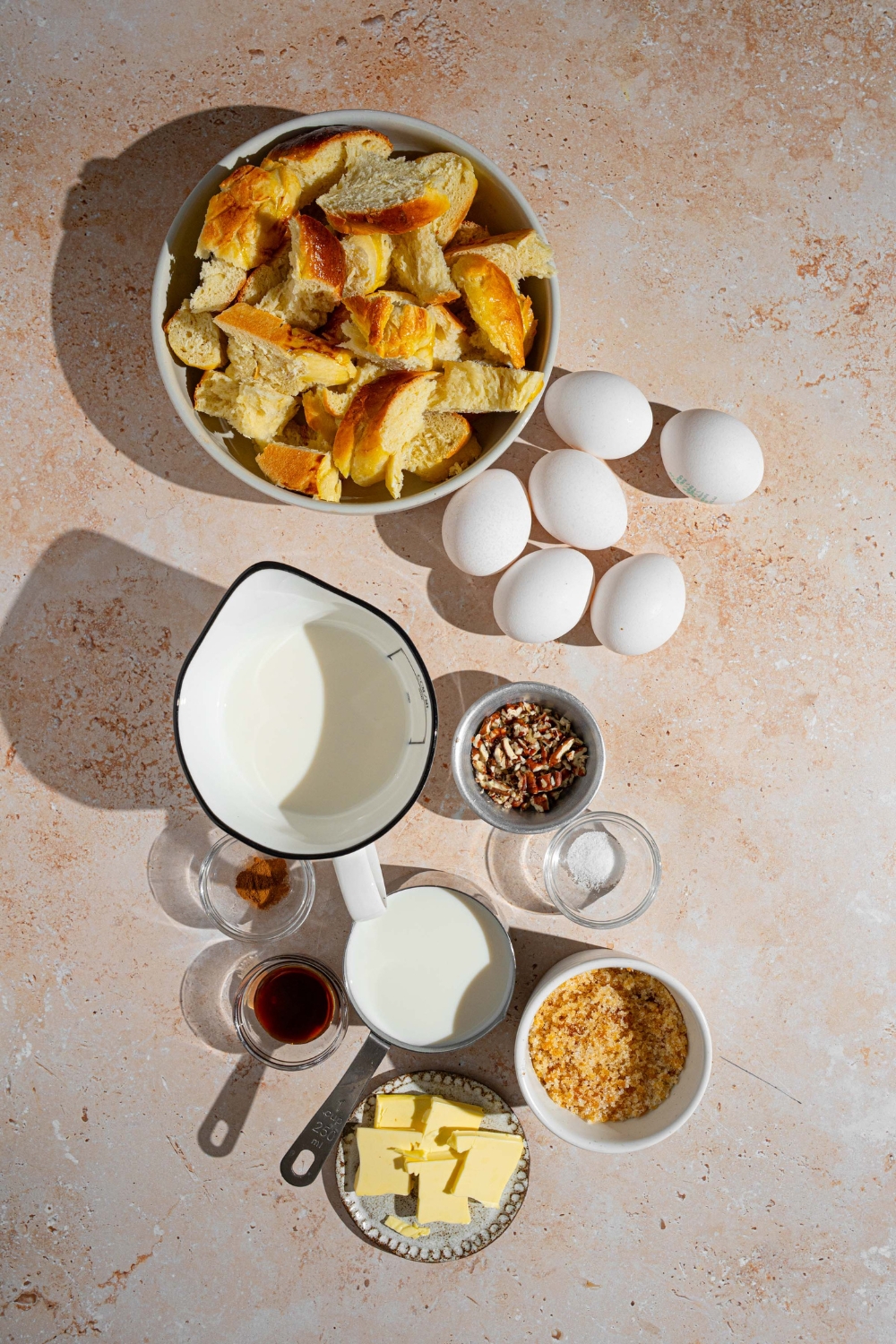An overhead shot of several bowls in various sizes containing ingredients to make challah french toast including challah bread, eggs, milk, heavy cream, butter, brown sugar, cinnamon, vanilla, salt, and pecans.
