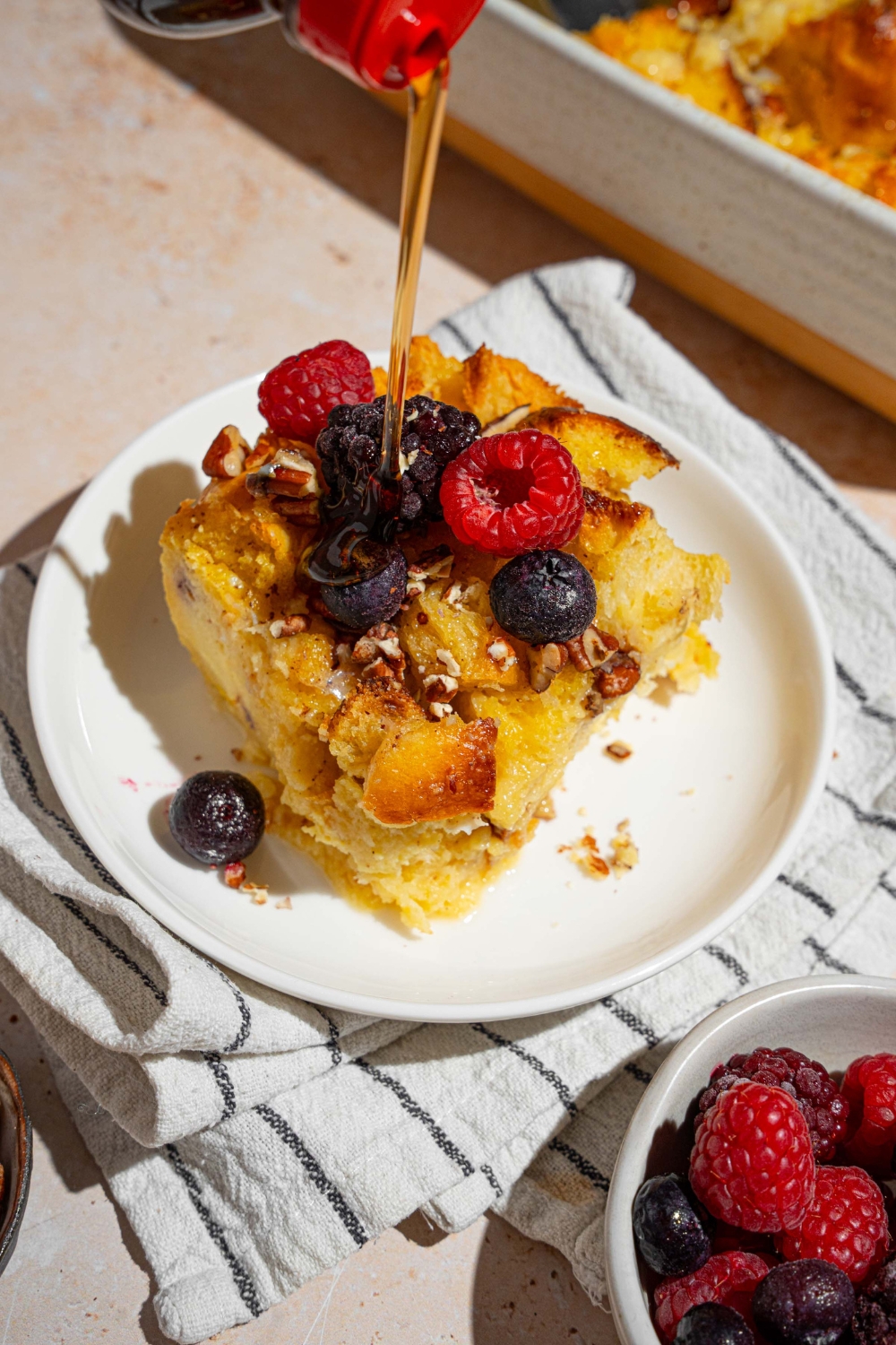 A white plate with a slice of challah french toast topped with mixed berries. Syrup is being poured on the plate. The plate is on a tan counter with a white striped napkin with a bowl of berries and baking dish of french toast.