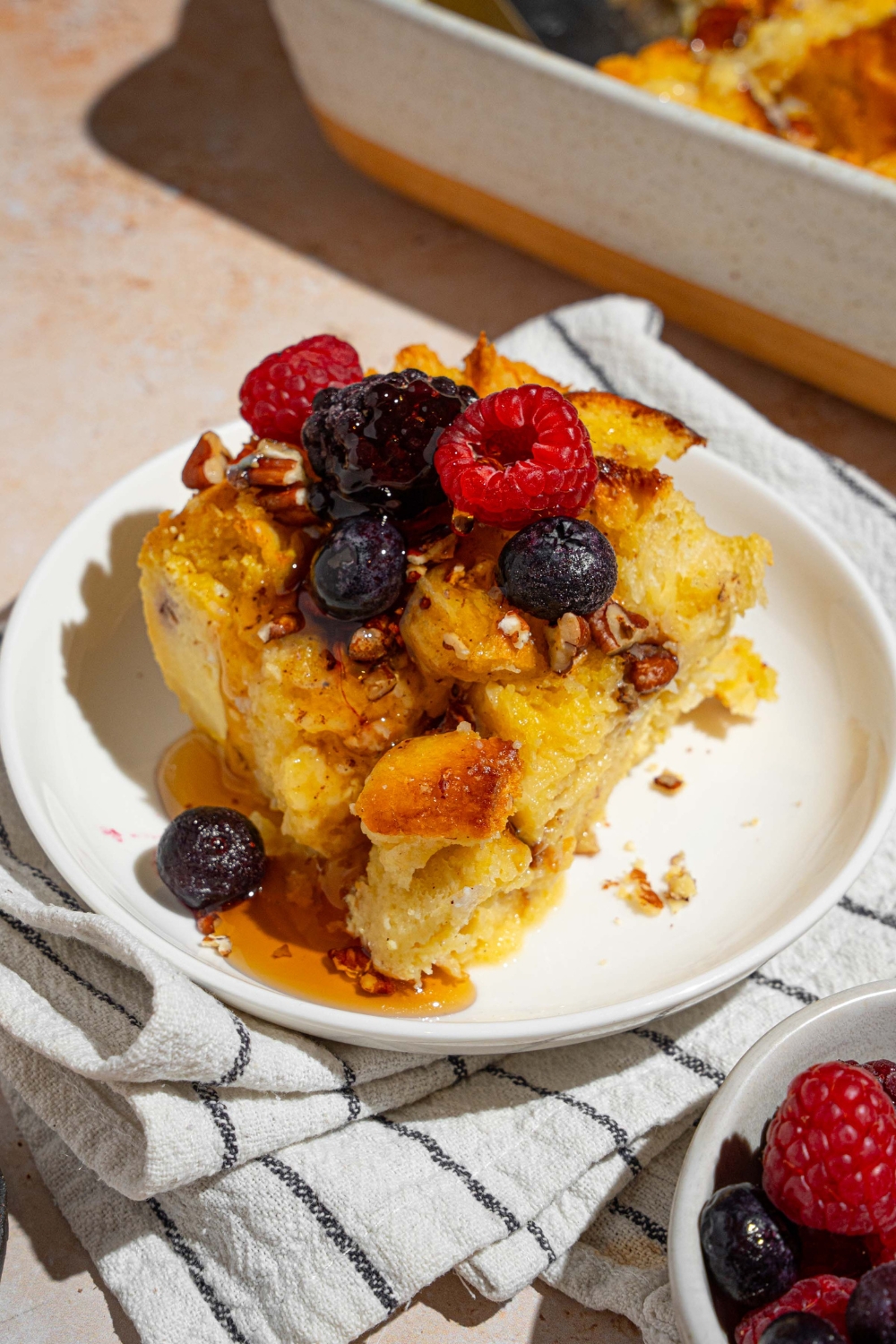 A white plate with a slice of challah french toast topped with mixed berries drizzled with syrup. The plate is on a tan counter with a white striped napkin with a bowl of berries and baking dish of french toast.