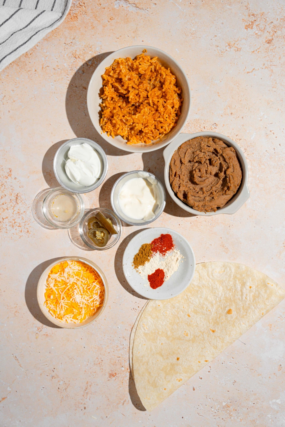 An overhead shot of several bowls in various sizes containing ingredients to make cheesy bean and rice burritos including tortillas, Spanish rice, refried beans, shredded cheese, mayo, sour cream, jalapenos, and seasonings.