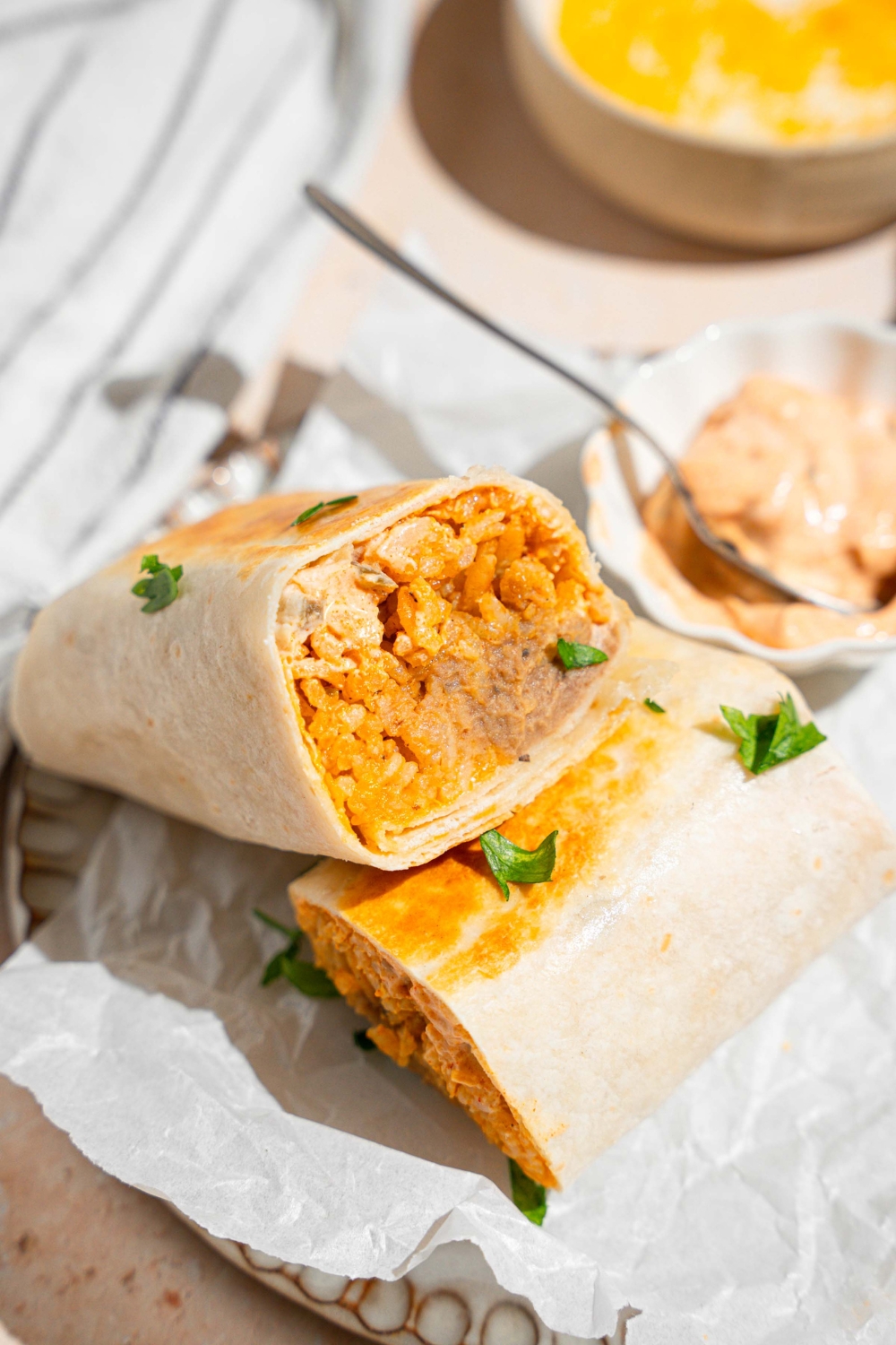 A cheesy bean and rice burrito sliced in half with each half stacked on the other. The burrito is on a plate lined with parchment paper with a small side of creamy jalapeno sauce with a spoon. The plate is garnished with fresh parsley. The plate is on a tan counter with small bowls of refried beans and shredded cheese.