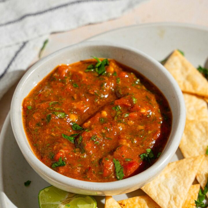 A bowl of Chipotle red chimichurri garnished with fresh parsley on a plate with tortilla chips. The plate is served with a lime wedge on a tan counter with a white striped napkin.