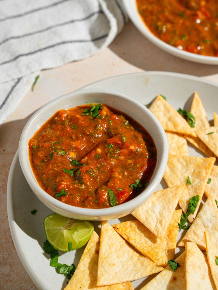 A bowl of Chipotle red chimichurri garnished with fresh parsley on a plate with tortilla chips. The plate is served with a lime wedge on a tan counter with a white striped napkin and bowl of chimichurri.