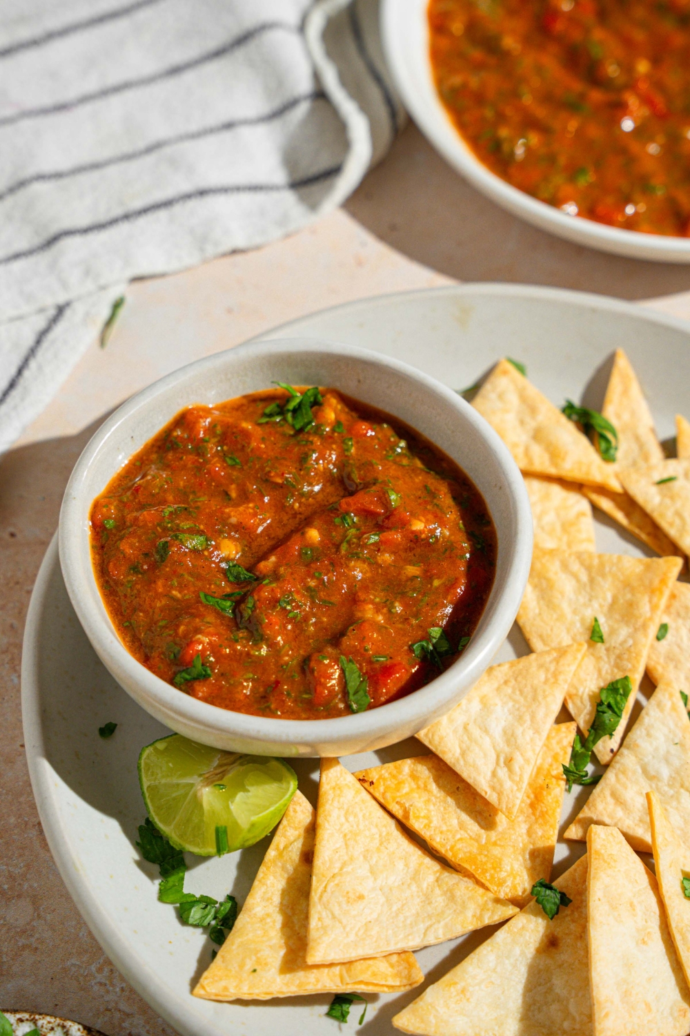 A bowl of Chipotle red chimichurri garnished with fresh parsley on a plate with tortilla chips. The plate is served with a lime wedge on a tan counter with a white striped napkin and bowl of chimichurri.