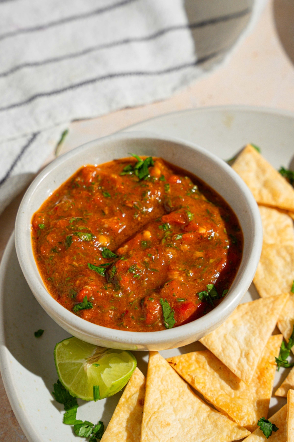 A bowl of Chipotle red chimichurri garnished with fresh parsley on a plate with tortilla chips. The plate is served with a lime wedge on a tan counter with a white striped napkin.