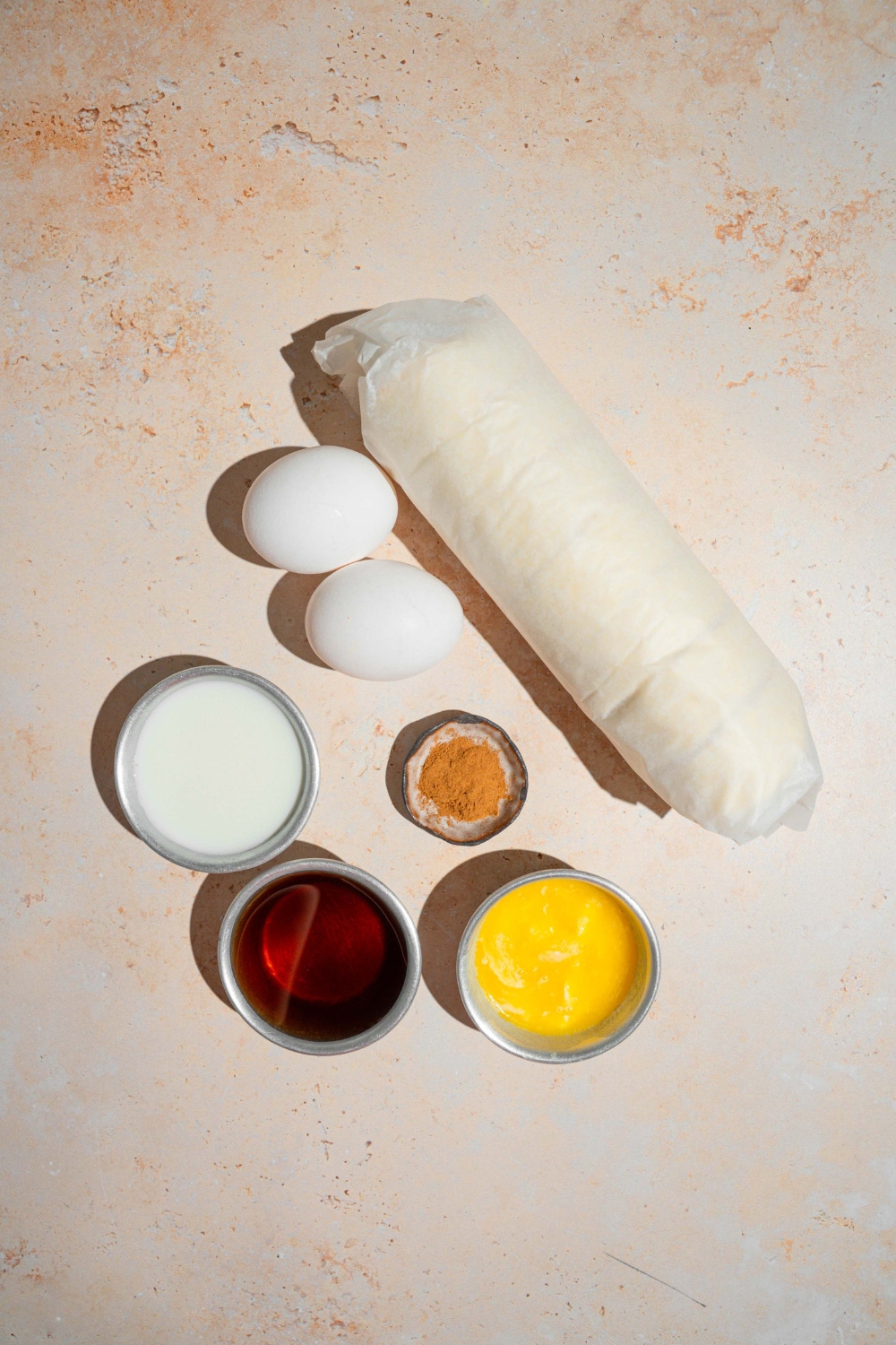 An overhead shot of several bowls in various sizes containing ingredients to make cinnamon roll french toast including a tube of cinnamon rolls, butter, eggs, cinnamon, vanilla, and milk.