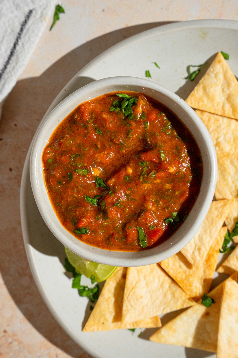 A bowl of Chipotle red chimichurri garnished with fresh parsley on a plate with tortilla chips. The plate is served with a lime wedge on a tan counter with a white striped napkin.