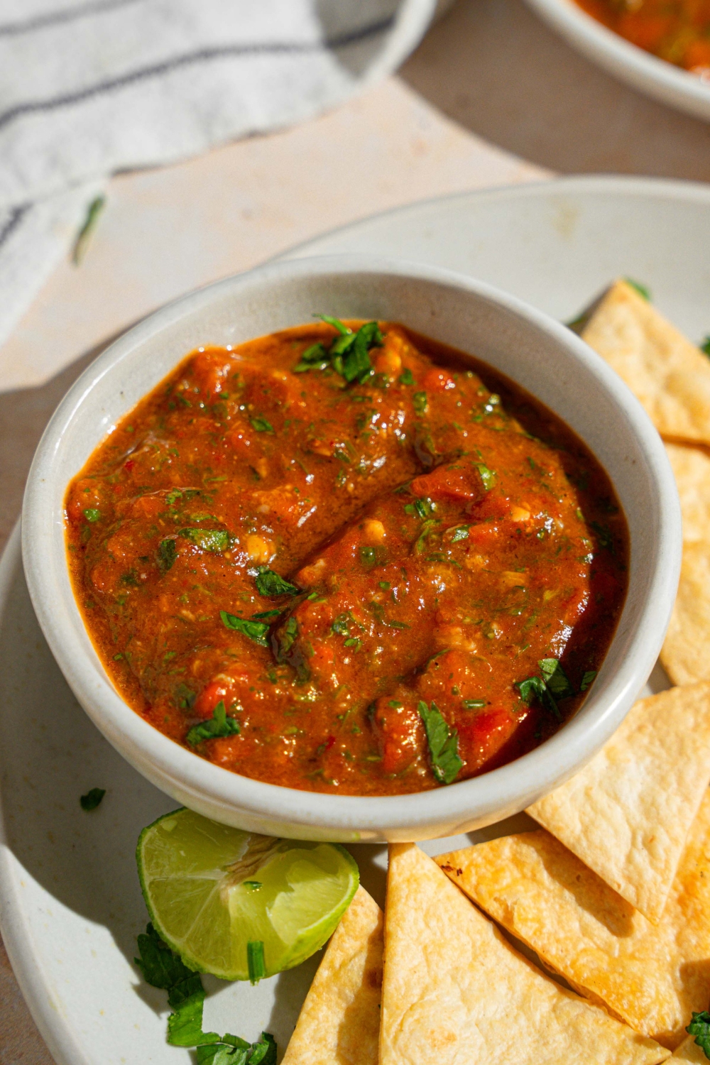 A bowl of Chipotle red chimichurri garnished with fresh parsley on a plate with tortilla chips. The plate is served with a lime wedge on a tan counter with a white striped napkin.