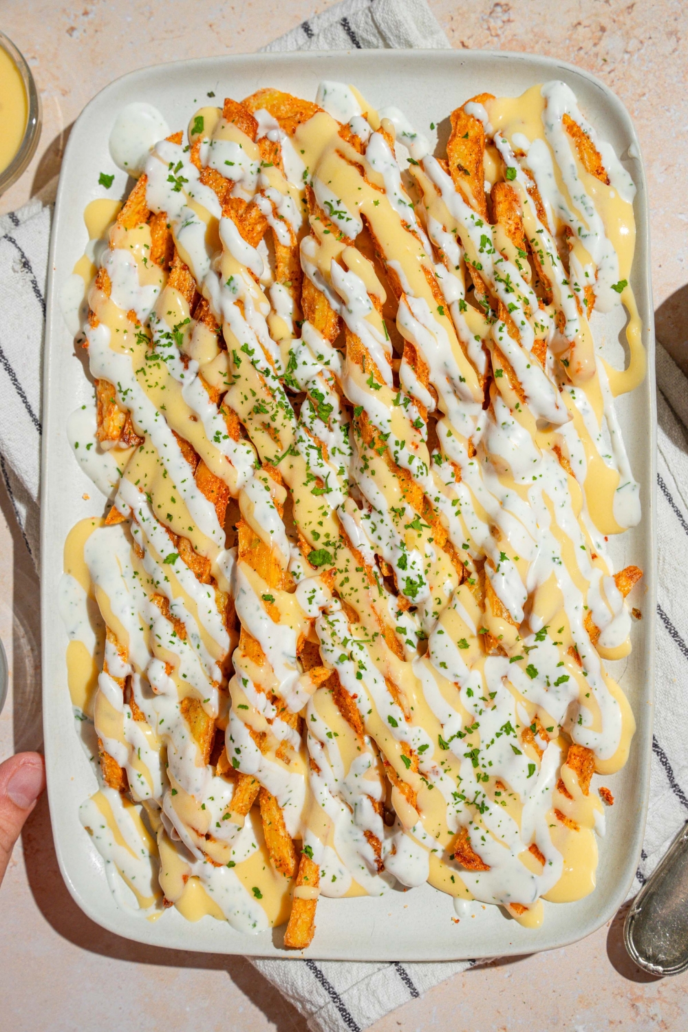 A white platter with copycat Wingstop Voodoo fries drizzled with cheese and ranch dressing. The plate is garnished with chopped parsley and served over a white striped napkin.