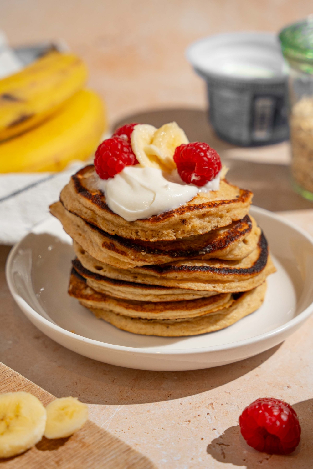 A white plate with a stack of cottage cheese banana pancakes topped with whipped cream, sliced banana, and raspberries. The plate is on a tan counter with bananas and a bowl of raspberries.
