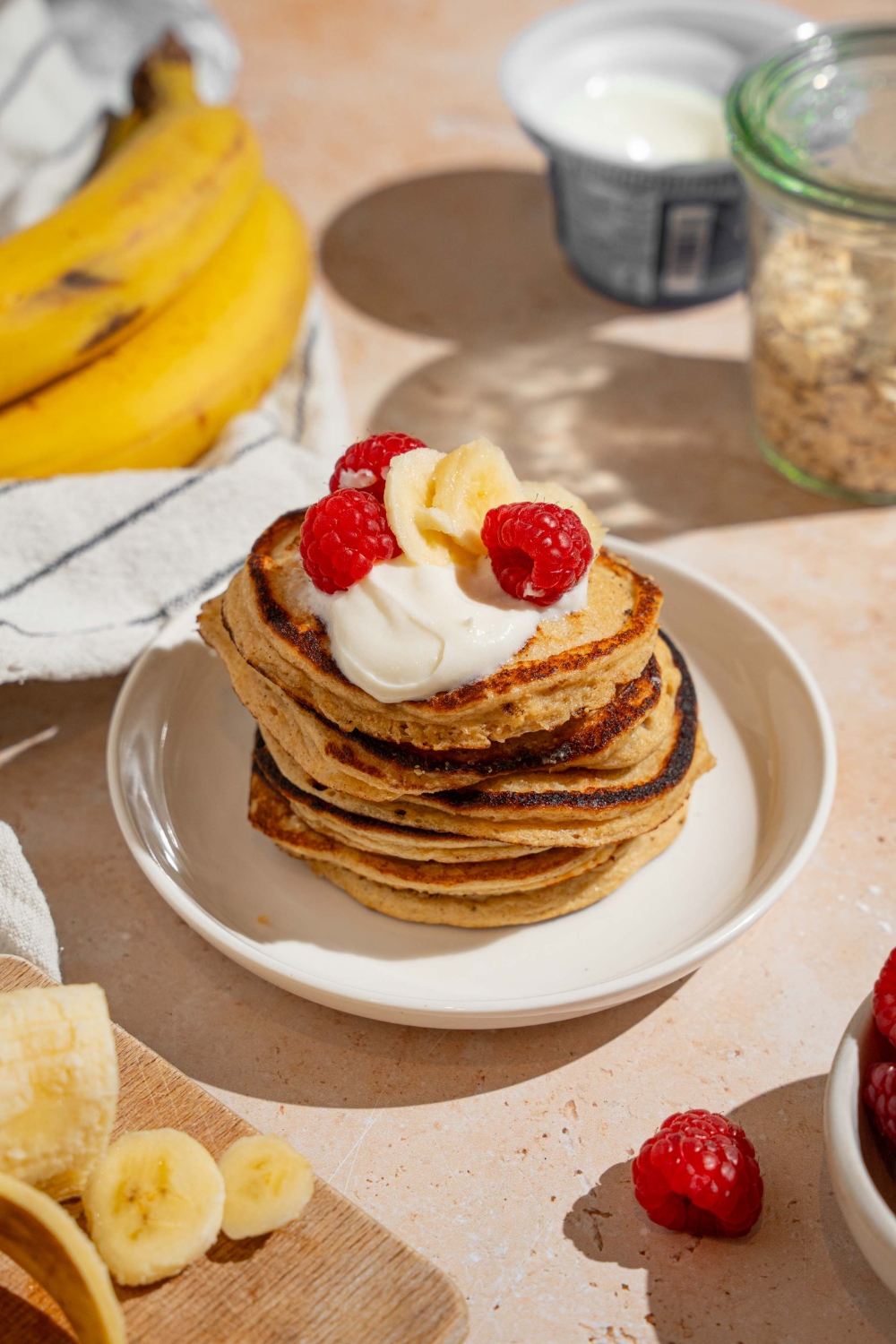 A white plate with a stack of cottage cheese banana pancakes topped with whipped cream, sliced banana, and raspberries. The plate is on a tan counter with bananas and a bowl of raspberries.