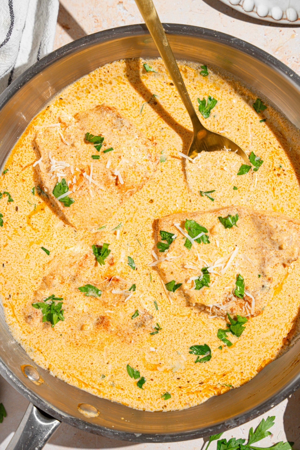 A skillet with a spoon stirring chicken breasts cooked in cajun alfredo sauce garnished with fresh parsley. The skillet is on a tan counter with a white striped napkin.