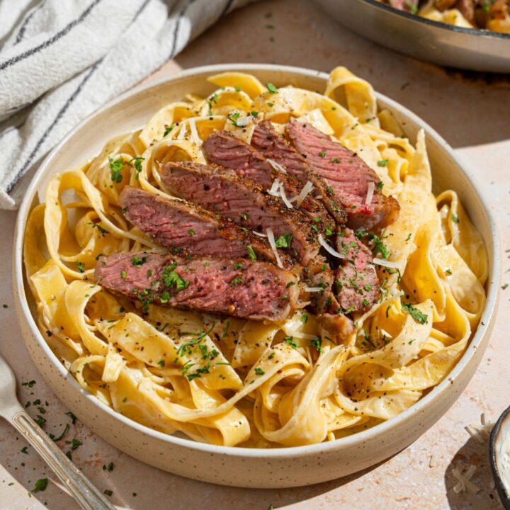 Fettuccine alfredo topped with sliced steak garnished with fresh parsley and grated cheese in a white bowl. The bowl is on a tan counter with a fork and white striped napkin.